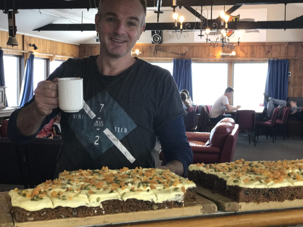 Member holding a coffee mug beside trays of carrot cake in the lodge lounge